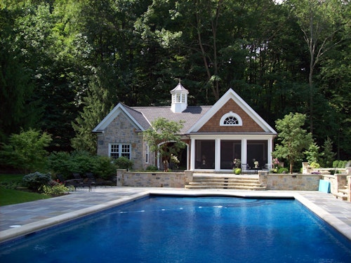 A serene backyard scene featuring a large rectangular swimming pool with clear blue water in the foreground. Beyond the pool, a charming stone pool house with a gabled roof and cupola is nestled among lush greenery. Stone steps lead to the entrance, flanked by potted plants and flowers, with stone tiles surrounding the pool area.