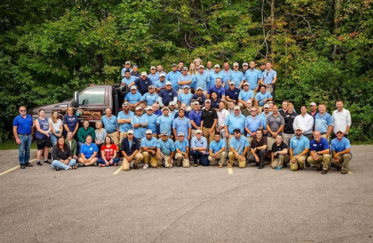 A large group of The Pattie Group employees poses for a team photo in front of a branded company truck, set against a backdrop of green trees. The team includes people in uniforms and casual wear, representing various departments and roles within the landscaping company.