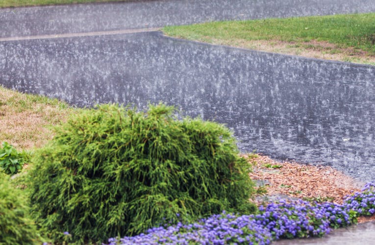 Residential yard drainage system installation in progress, featuring gravel-filled trenches with green catch basins and surrounding mounds of excavated soil. A skid steer and two workers are visible in the background near a house with a deck.